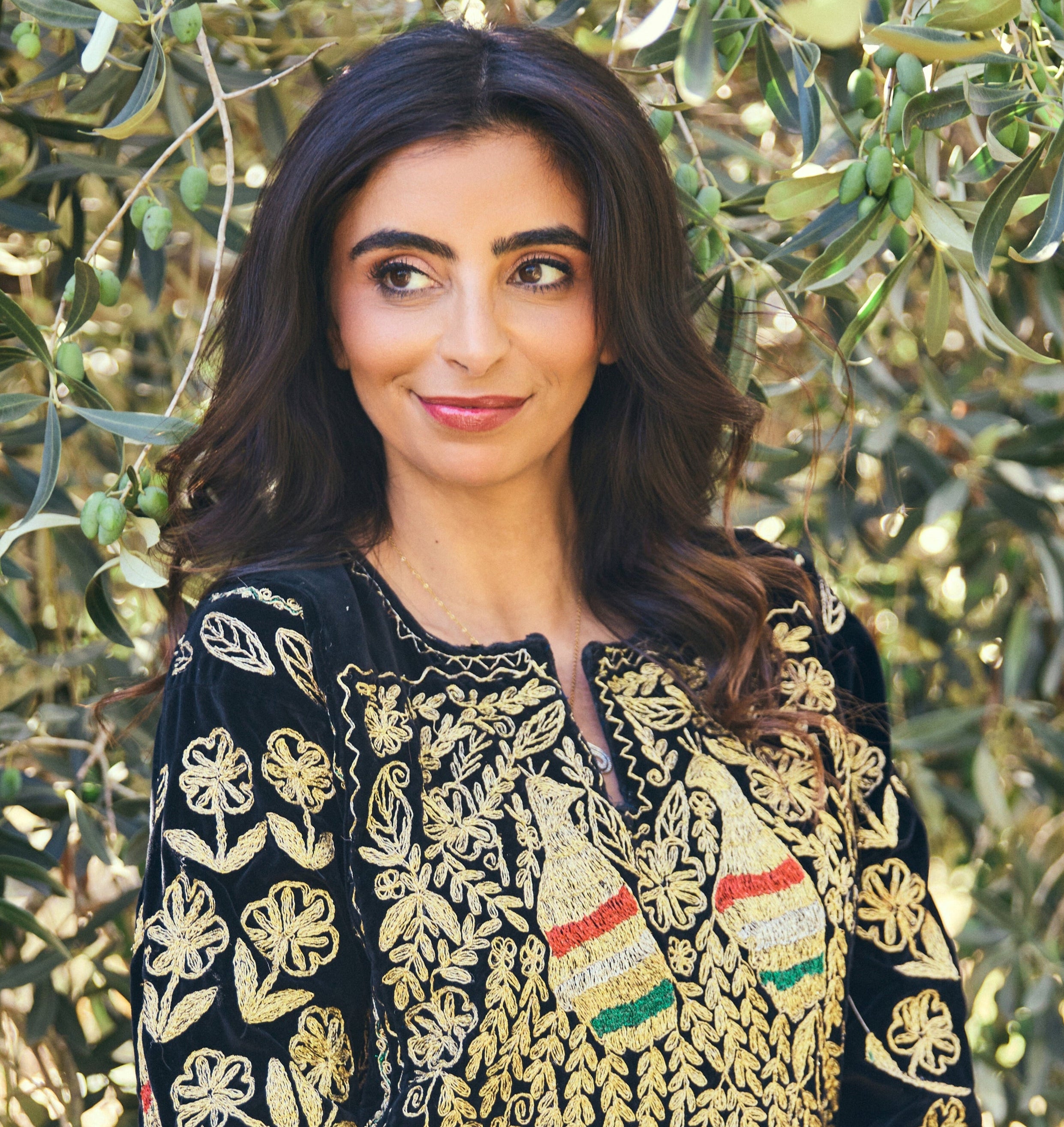 Woman wearing a black and gold Palestinian embroidered traditional outfit with green foliage in the background