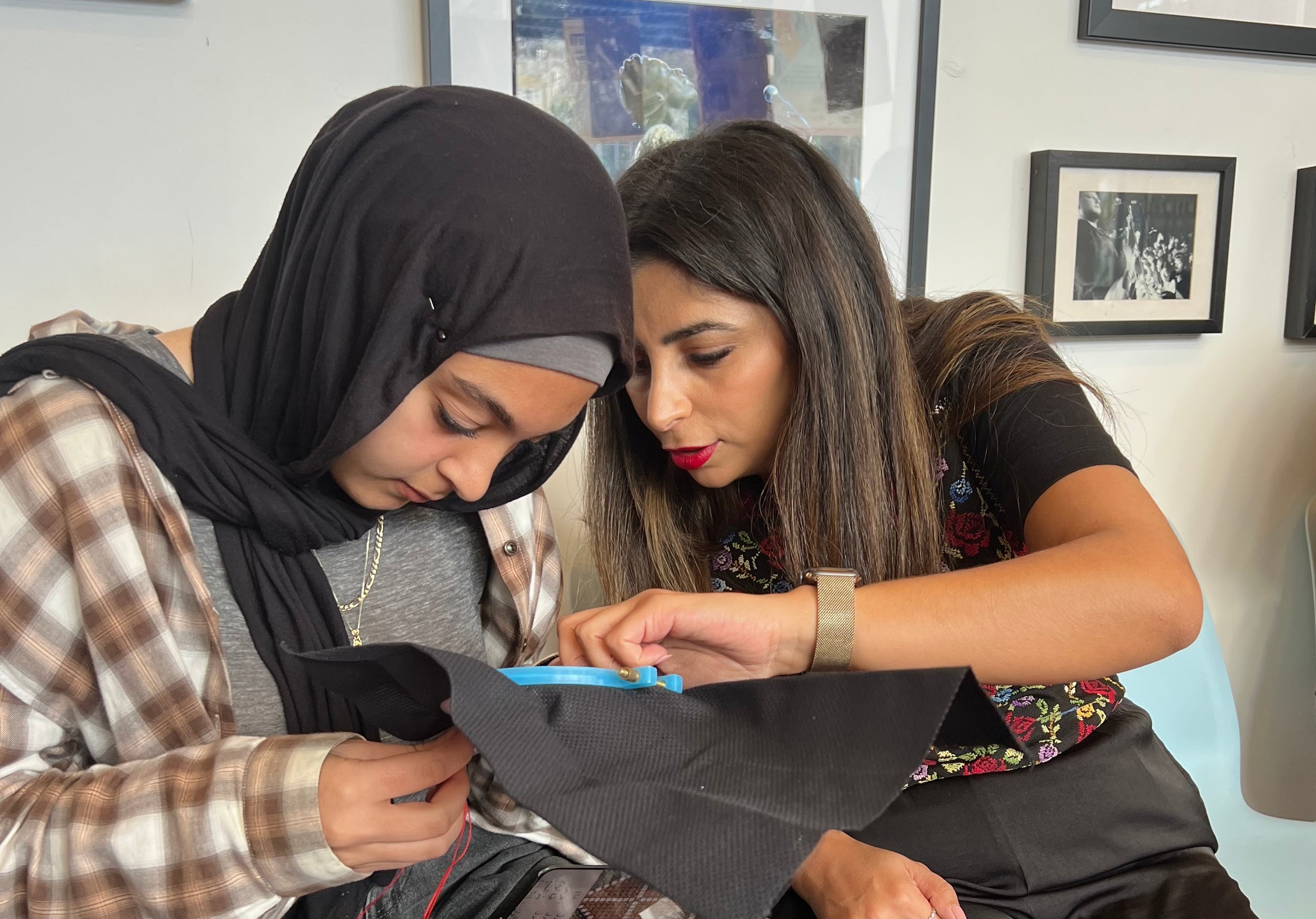 Two women seated indoors, one wearing a black hijab and plaid shirt while the other assists with embroidering a piece of black fabric.